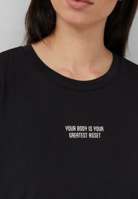 Schwarzes Baumwoll-T-Shirt mit weißem Text, der "DEIN KÖRPER IST DEIN GRÖSSTES ASSET" in einer modernen serifenlosen Schriftart zeigt.