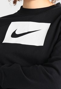 Svart sweatshirttopp med långa ärmar, prydd med ett stort vitt rektangulärt märke med en svart Nike-logga i mitten. Mjuk tygstruktur.