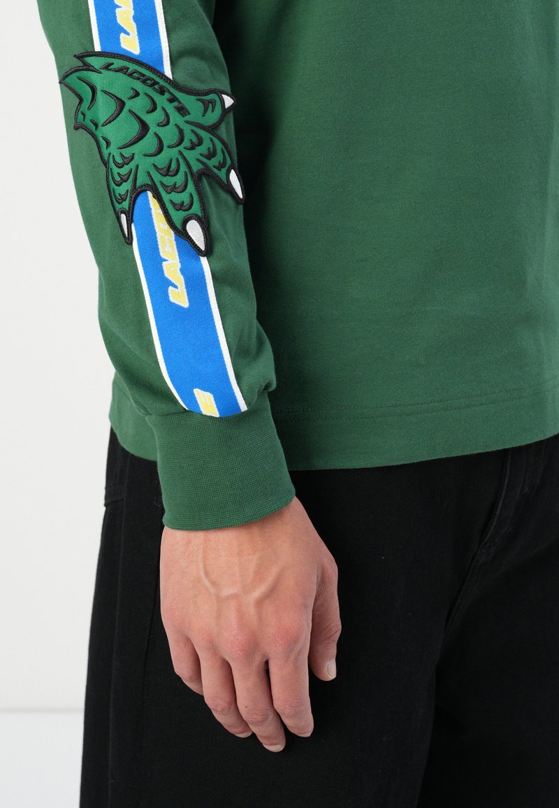 Zielona koszula z długim rękawem z haftowanym zielonym wzorem dłoni na rękawie, z niebieskimi i żółtymi paskami wzdłuż ramienia.