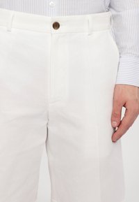Hvit linbukser med en naturlig tekstur, med frontknapp og sidelommer, vist med en hånd hvilende på låret.