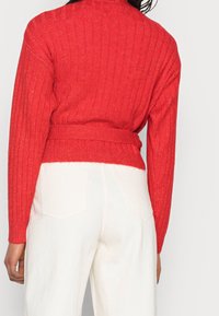 Suéter rojo de punto acanalado con cuello alto y cinturilla anudada, combinado con pantalones de pierna ancha blancos que tienen bolsillos traseros.