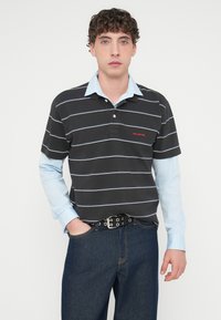 Πλεκτό μπλουζάκι πόλο με ρίγες σε ασπρόμαυρο χρώμα και ανοιχτό μπλε γιακά, συνδυασμένο με ανοιχτό μπλε μακρυμάνικο πουκάμισο από μέσα και σκούρο μπλε τζιν παντελόνι.