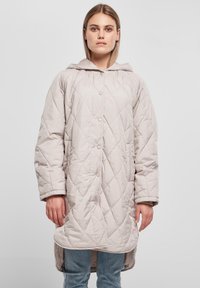 Lys grå quiltet lengre jakke med hette, med trykknapper og to sidelommer, paret med denimjeans.