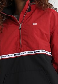 Žena ve červené a černé mikině s půlzipem, na které je pruh s logem "Tommy Jeans" přes hruď a malé logo na levé horní straně.
