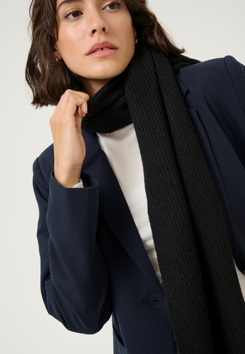 Scarf nero a costine realizzato in morbido tessuto, indossato sopra una maglietta bianca e un blazer blu navy, con un focus ravvicinato sulla texture e sui dettagli.