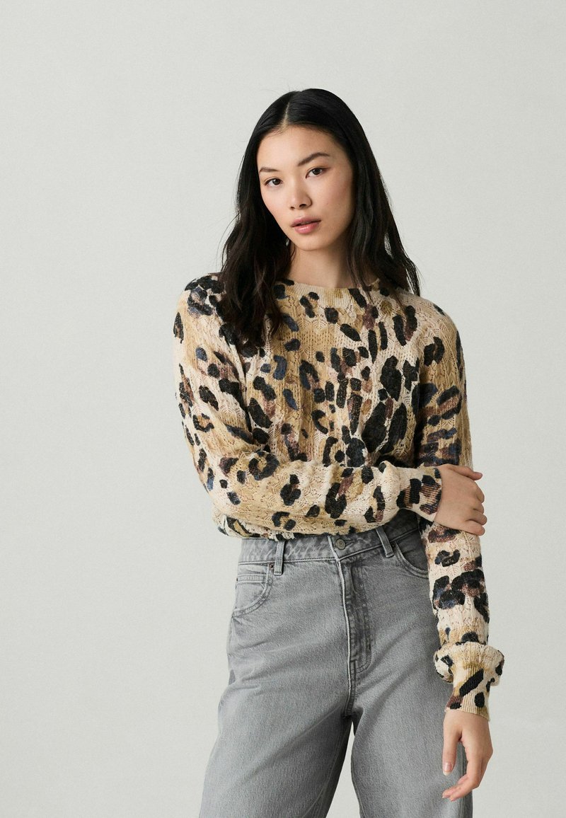 Beżowy sweter w leopardzie w czarne i brązowe plamy, z szerokim dekoltem i długimi rękawami, w zestawieniu z szarymi spodniami z wysokim stanem.