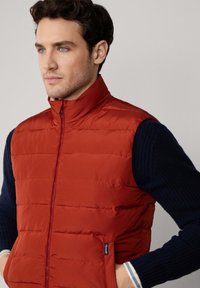 Chaleco acolchado rojo con cuello alto, con costuras verticales y cremallera frontal. Lleva puesto un suéter de punto texturizado azul oscuro.