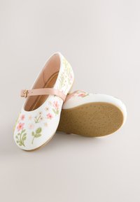 Chaussures Mary Jane blanches brodées de fleurs avec bride rose et semelle beige embossée de motifs papillon et étoile.