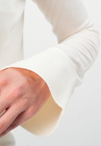 Hvit skjorte med lange ermer som har en ribbet tekstur og brede, scallop-formede mansjetter. Mansjettene har lett utfransede kanter, noe som gir designet ekstra detalj.