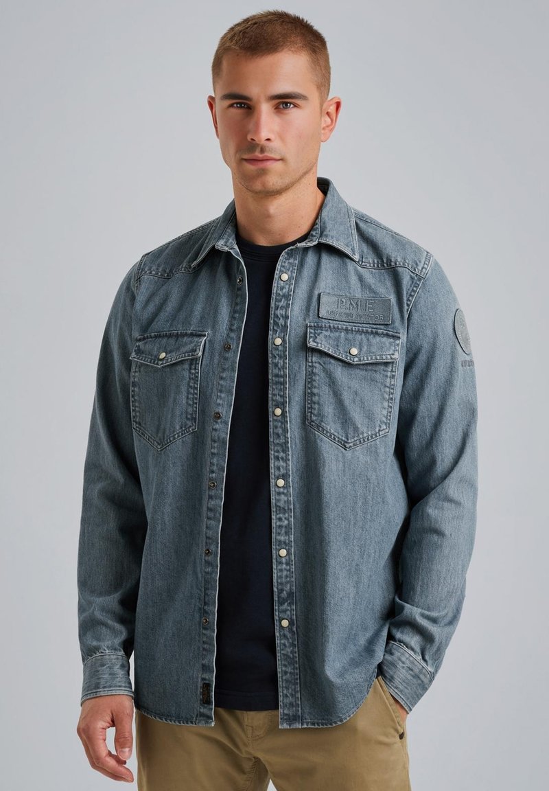 Jasnoniebieska koszula dżinsowa z długimi rękawami, dwoma kieszeniami na piersi, zapięciem na guziki z przodu i naszywką po lewej stronie. Noszona na ciemnej bluzce.