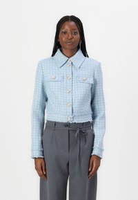 Vaaleansininen tweed-tyylinen lyhyempi takki, joka sisältää kauluksen, etutaskut ja suuret koristepainikkeet, joissa on rispaantuneet helmapitsit.