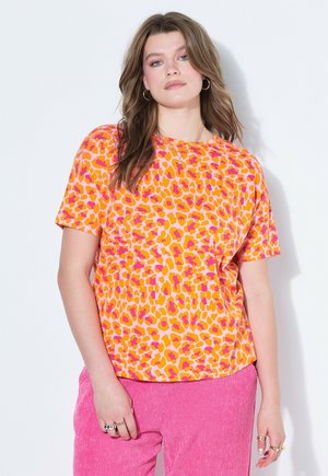 Frau mit welligem braunem Haar trägt ein leuchtend orange-rosa kurzärmeliges Leopardenmuster-Shirt und texturierte rosa Hose vor weißem Hintergrund.