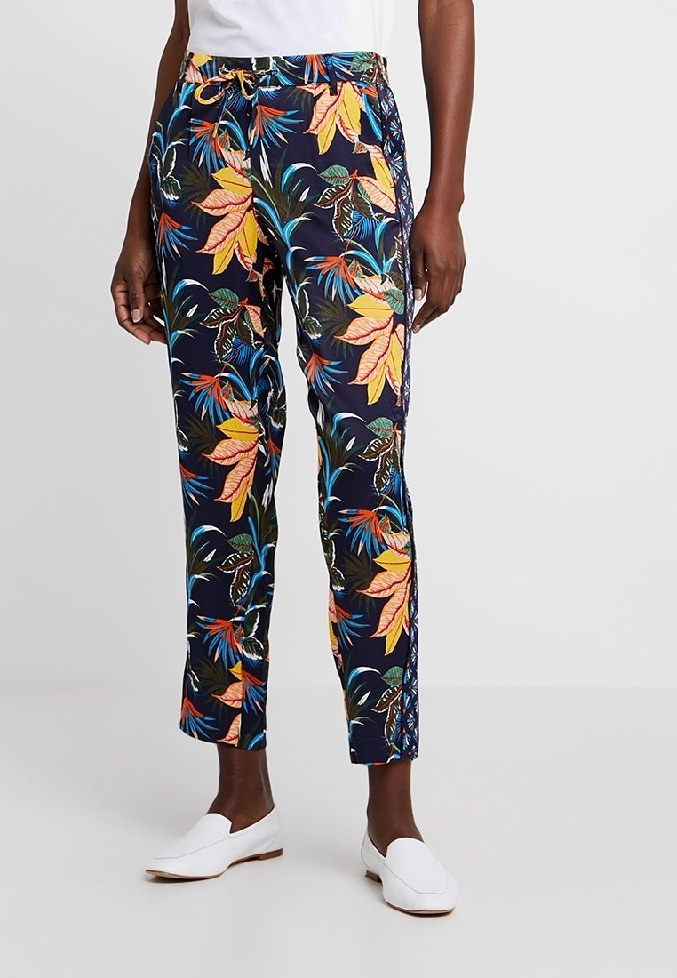 Personne portant un pantalon bleu marine avec des motifs de feuilles tropicales colorées et des chaussures blanches à enfiler, debout devant un fond clair uni.
