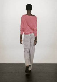 Jasnoróżowy sweter z opuszczonymi ramionami, luźny krój, w połączeniu z jasnoszarymi szerokimi dżinsami z widocznymi przeszyciami i kieszeniami, na modelce widzianej od tyłu.