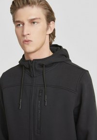 Felpa nera con zip e finitura testurizzata. Presenta un cappuccio con coulisse, una tasca sul petto con zip e una branding discreta sulla manica.