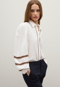 Camisa blanca de manga larga con acentos bordados en marrón en las mangas, que presenta un cierre de botones en la parte delantera y un cuello. Combinada con pantalones azul marino.