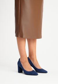 Des chaussures à talons hauts en daim bleu avec un bout pointu et un talon épais, assorties à une jupe en cuir marron. Texture lisse et lignes de design nettes.