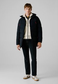 Czarna kurtka puchowa z czerwonymi akcentami, zapinana na zamek, z kapturem, na kremowej warstwie polarowej. Stylizowana z ciemnymi dżinsami i białymi trampkami.