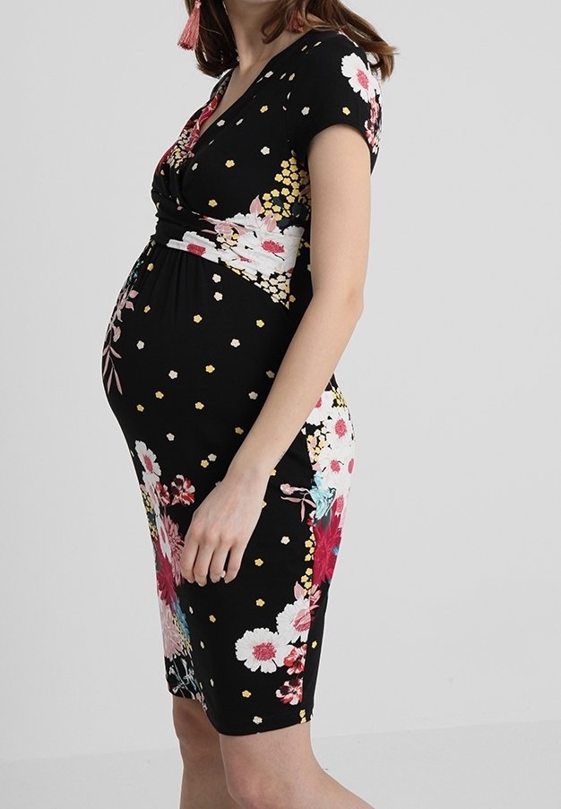 Femme enceinte portant une robe noire fleurie mi-longue à manches courtes, debout de profil sur un fond uni.