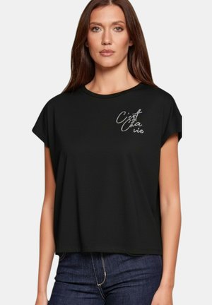 Donna che indossa una maglietta nera a maniche corte con la scritta "C'est Ça la vie" sul lato sinistro del petto, abbinata a jeans blu scuro, in piedi davanti a uno sfondo semplice.