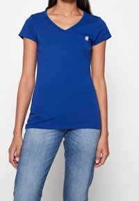 Kvinna iklädd en figursydd blå V-ringad kortärmad T-shirt med en liten vit logga på bröstet, tillsammans med ljusblå denimjeans.