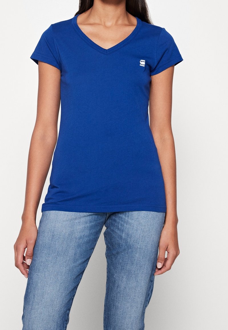 Kvinna iklädd en figursydd blå V-ringad kortärmad T-shirt med en liten vit logga på bröstet, tillsammans med ljusblå denimjeans.