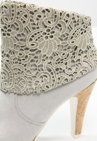 Gray suede ankle boot with a decorative light gray lace trim and a cork heel. The boot has a rounded toe and smooth texture.