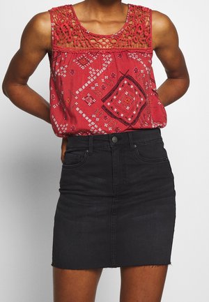Vrouw draagt een rood topje zonder mouwen met patroon en haakwerkdetails, gecombineerd met een zwarte denim minirok met hoge taille.