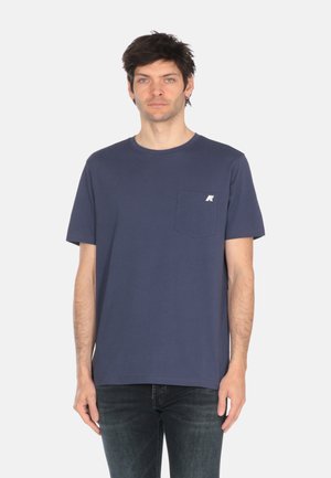 Uomo con capelli corti e scuri che indossa una maglietta blu navy a maniche corte con un piccolo logo bianco sulla tasca del petto, in piedi davanti a uno sfondo bianco.