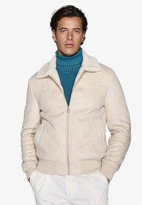 Beige ruskinds bomberjakke med en blød krave, frontlynlås og ribbede manchetter. Bæres over en struktureret teal turtleneck sweater.