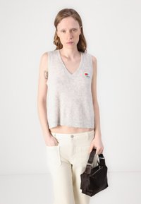 Top de punto sin mangas en gris claro con escote en V, que presenta un parche con el logo. Combinado con pantalones de pierna ancha en color crema y un pequeño bolso oscuro.