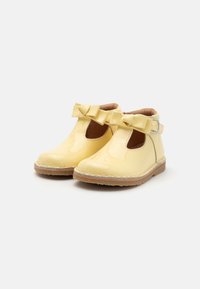 Żółte lakierowane buty z okrągłym noskiem, zapinane w stylu T z dużymi kokardami z przodu. Posiadają zapięcie na klamrę i teksturowaną podeszwę.
