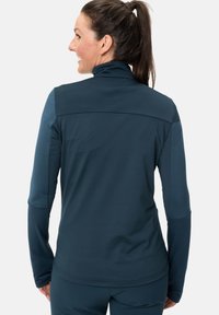 Giacca sportiva teal realizzata in tessuto elasticizzato, con colletto alto e spalle senza cuciture, progettata per comfort e mobilità.