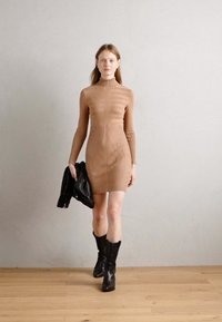 Robe moulante à manches longues en côtes couleur beige, dotée d'un col montant et d'un motif rayé subtil. Portée avec une veste en cuir noir et des bottes jusqu'aux genoux.