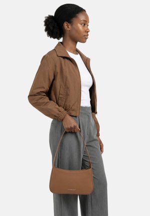 Junge Frau in brauner Jacke und grauer Hose hält eine braune Handtasche und schaut nach rechts vor einem einfarbigen Hintergrund.