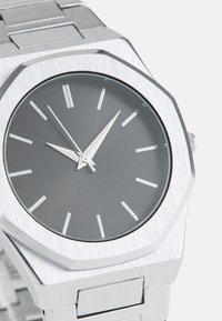 Sølv octagonal ur med et børstet metalarmbånd. Grå urskive med minimalistiske timemarkører og sølvfarvede visere. Rent, moderne design.