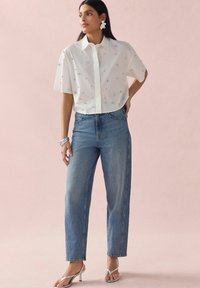 Chemise blanche à manches courtes avec des embellissements argentés, associée à un jean taille haute bleu clair et des sandales à talons argentées. Fond rose.