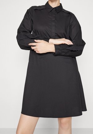 Osoba nosząca czarną sukienkę koszulową z długim rękawem i koronkową lamówką na rękawach, z założonymi na wysokości talii ramionami, na jednolitym jasnym tle.