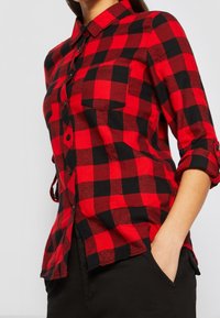 Röd- och svartrutig flanellskjorta med krage, uppvikt ärm och en framficka. Tyget verkar mjukt med en texturerad yta.