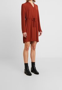 Rustovärinen mekko, jossa on pitkät hihat, V-halkio, vyötä ympäröivä vyötärö ja sivuhalkiot, paritettuna mustien nilkkasaappaiden kanssa, jotka ovat saanut tyylikkään viimeistelyn.