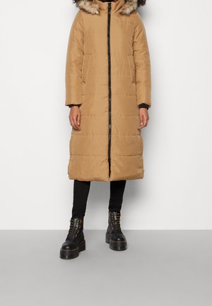 Personne portant un long manteau matelassé beige avec capuche bordée de fourrure, des leggings noirs et des bottes à plateformes noires à lacets, debout sur un sol gris.