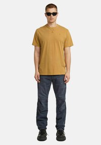 T-shirt a maniche corte giallo senape con scollo rotondo e dettagli di cucitura, abbinato a pantaloni cargo blu scuro e sneakers nere.