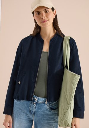 Giovane donna che indossa un cappellino beige chiaro, giacca blu navy con zip, canotta verde e jeans blu, che porta una borsa trapuntata verde chiaro sulla spalla.