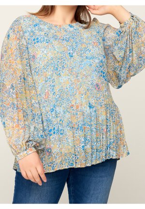 Woman wearing a loose, long-sleeve floral blouse with blue, yellow, and orange hues paired with blue jeans.