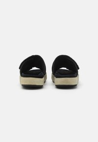 Sandali in suede nero con suola testurizzata beige, apertura sul retro e dettagli della cinturino regolabile per una calzata sicura. Design piatto per il massimo comfort.