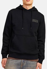 Mężczyzna w czarnej bluzie z kapturem z przednią kieszenią i logo "JACK & JONES" na piersi, w połączeniu z niebieskimi dżinsami i brązową koszulą.