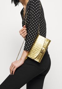 Kultaisesta keinonahasta valmistettu olkalaukku krokotiilikuosisella pinnalla, ketjuhihnalla ja etupuolella kannella. Kompakti ja suorakulmainen muoto.