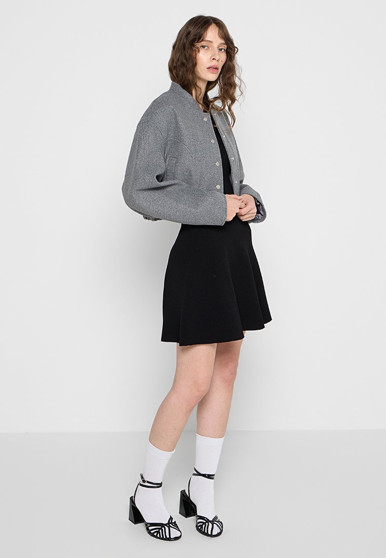 Veste grise courte avec col arrondi et fermeture à boutons, portée sur une robe noire. Les accessoires comprennent des chaussettes blanches et des sandales à talons noires.