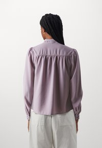 Light purple blouse with puffed sleeves and a buttoned cuff, featuring a gathered back detail and a smooth texture.