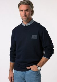 Sweatshirt marine avec des poignets et un col côtelés, présentant un patch en cuir carré avec le logo 'BOSTON'. En dessous, un col de chemise à carreaux bleu est visible.